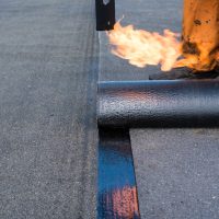 Roofer man worker at building site installing roll of roofing felt with gas blowpipe torch during construction works. flame during welding of a waterproofing membrane on a roof